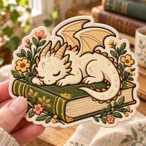 May include: An embroidered patch featuring a cream-colored dragon sleeping on a green book. The design includes yellow wings, brown horns, and surrounding floral elements in shades of pink, yellow, and green with dark brown outlines.
