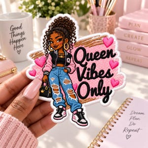 May include: A decorative embroidered patch featuring a Black girl with curly hair wearing a black and pink varsity jacket, blue distressed jeans, and sneakers. The patch includes the black text Queen Vibes Only and several small pink hearts.