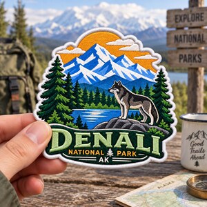 May include: An embroidered patch featuring a gray and black wolf standing on a rock before a blue lake, green pine trees, and snow-capped mountains under an orange sky. The text reads DENALI NATIONAL PARK AK with a small pine tree icon.