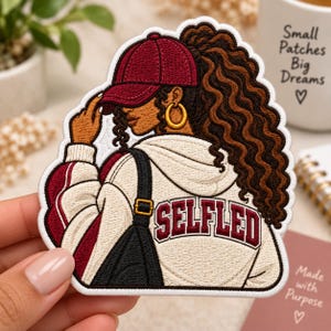 May include: An embroidered patch featuring a person with long curly brown hair wearing a maroon baseball cap, gold hoop earrings, and a cream-colored hoodie with maroon accents. The back of the hoodie displays the text SELFLED in maroon block letters.