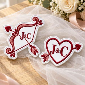 May include: Two white embroidered patches with red and pink accents. One patch is shaped like a bow and arrow, the other like a heart pierced by an arrow. Both feature the embroidered letters J&C in a dark red serif font.