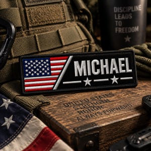 May include: A rectangular embroidered patch featuring a black background with a diagonal split. The left side displays the American flag in red, white, and blue. The right side features the white embroidered name MICHAEL above two white stars.