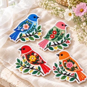 May include: Four embroidered bird patches featuring vibrant colors and floral patterns. Each bird is perched on a branch with green leaves and small berries. The birds are blue, pink, red, and orange, each decorated with intricate flower designs.