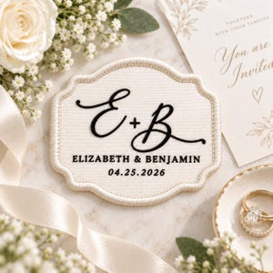 May include: A cream-colored fabric patch with a scalloped edge featuring the black embroidered initials E + B, the names ELIZABETH & BENJAMIN, and the date 04.25.2026. The patch is displayed alongside white flowers, a ribbon, and a wedding invitation.