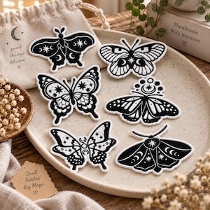 May include: Six embroidered patches featuring black and white butterfly and moth designs with celestial motifs including stars and crescent moons. The patches are arranged on a round ceramic plate alongside a small beige fabric drawstring bag.