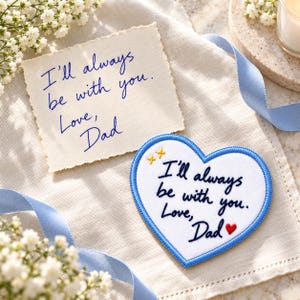 May include: A white heart-shaped fabric patch with a blue embroidered border featuring the dark blue stitched text I'll always be with you. Love, Dad and a small red heart. Next to it is a paper note with the same handwritten message in blue ink.