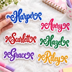 May include: Six embroidered name patches featuring the names Harper in blue, Avery in pink, Scarlett in red, Hazel in green, Grace in purple, and Riley in yellow. Each name is written in a cursive script and flanked by two small bow icons.