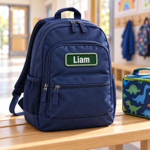 May include: A navy blue backpack featuring a rectangular green patch with the white text Liam embroidered on the front. The bag includes a top handle, adjustable shoulder straps, a front zippered pocket, and side mesh pockets for storage.