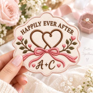 May include: An embroidered patch with a cream-colored background and a scalloped edge. It features the text "HAPPILY EVER AFTER" in brown, two interlocking brown hearts, a pink ribbon, green leaves, small pink hearts, and "A + C" in brown.