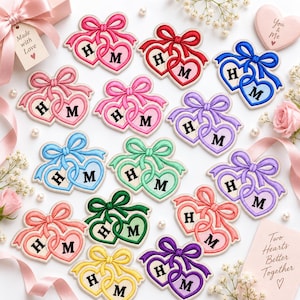 May include: A collection of embroidered heart-shaped patches featuring interlocking hearts tied with a bow. Each patch displays the letters H and M in black. The patches come in various colors including pink, red, blue, purple, green, and yellow.