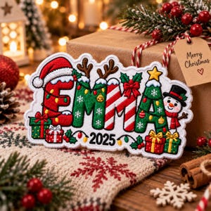 May include: A white embroidered Christmas ornament shaped like the name EMMA with the year 2025. The letters feature red, green, and candy cane patterns, decorated with a Santa hat, reindeer antlers, a snowman, gifts, and holiday lights.