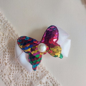 Puede incluir: Un accesorio para el cabello con una base de lazo de cinta blanca rematada con una forma de mariposa cubierta de lentejuelas iridiscentes multicolores. Una perla artificial blanca, redonda y lisa, está centrada en la mariposa.