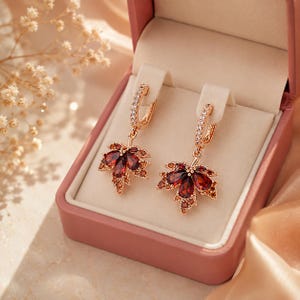May include: A pair of rose gold hoop earrings featuring a dangling maple leaf charm. Each leaf is encrusted with deep red pear-shaped gemstones and smaller orange accent stones. The hoop portion is lined with tiny clear white crystals.