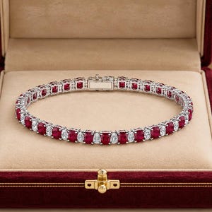 Pode incluir: Uma pulseira de ténis com rubis vermelhos de corte oval e diamantes brancos transparentes de corte redondo alternados numa banda de metal prateado. A pulseira inclui um fecho retangular e é apresentada numa caixa de joias forrada.