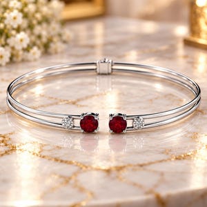 Puede incluir: Una pulsera abierta de tono plateado con dos piedras redondas de color rojo intenso en el centro, flanqueadas por dos piedras redondas transparentes más pequeñas a cada lado. La pulsera consta de dos bandas paralelas con una bisagra trasera.