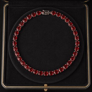May include: A circular tennis necklace featuring a continuous row of faceted, deep red oval-cut stones set in a silver-toned metal frame. The necklace includes a secure metal box clasp closure and is displayed in a black velvet-lined jewelry box.