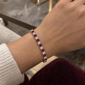 Può includere: Un bracciale tennis caratterizzato da un motivo ripetuto di rubini rossi a taglio rotondo e diamanti bianchi trasparenti incastonati in una fascia di metallo color argento. Le pietre sono disposte in fila, alternando gemme rosse e bianche.