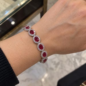 Puede incluir: Una pulsera de eslabones en tono plateado que presenta una serie de rubíes de color rojo intenso con forma ovalada. Cada rubí está rodeado por un halo de pequeñas piedras claras y brillantes engastadas en un marco metálico.