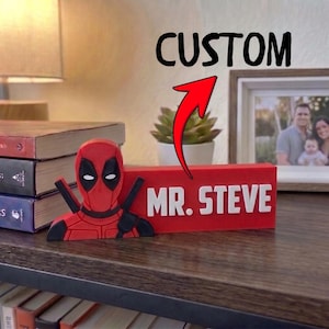 May include: A red rectangular desk sign featuring a 3D Deadpool character figure on the left side and the white text MR. STEVE on the right. Above the sign, a red arrow points to the text CUSTOM, indicating a personalized name plate.