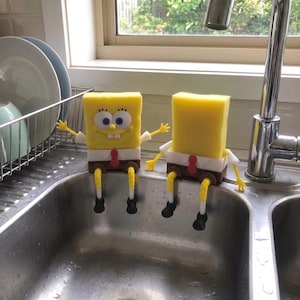 Op de afbeelding: Twee gele rechthoekige sponzen in de vorm van Spongebob Squarepants, elk met een wit overhemd, rode stropdas, bruine broek en gele ledematen met zwarte schoenen. De ene spons heeft een lachend gezicht met blauwe ogen, de andere is blanco.