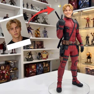 May include: A collectible action figure of a character in a red and black tactical suit with a utility belt, katana swords on the back, and a head sculpt featuring blonde hair. A separate photograph of the same person is displayed to the left.