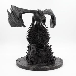 以下が含まれることがあります： マットブラックの3Dプリントフィギュア。翼を広げたドラゴンが、無数の剣の刃で構成された玉座の上に鎮座している。全体は円形の台座に取り付けられており、ドラゴンの鱗の複雑なディテールと、剣の玉座のギザギザした鋭いエッジが表現されている。