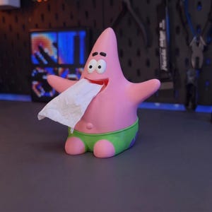 Op de afbeelding: Een roze, stervormige tissuehouder met een wijd open mond waar een witte tissue uitsteekt. Het figuur heeft twee grote ogen, dunne zwarte wenkbrauwen en draagt een groene broek met paarse bloemmotieven aan de zijkanten.