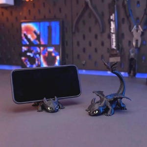 Puede incluir: Dos figuras de dragón negras con ojos grandes y orejas puntiagudas. Una figura actúa como soporte horizontal para smartphone, sosteniendo un dispositivo móvil rectangular negro. La segunda figura está colocada a la derecha, de pie sobre sus cuatro patas con la cola curvada hacia arriba, mostrando alas detalladas y un cuerpo texturizado.