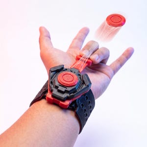 May include: A black and red wrist-mounted toy featuring a textured strap and a central mechanical housing. A red circular projectile is shown mid-air, connected to the device by a thin, translucent filament extending from the main unit.