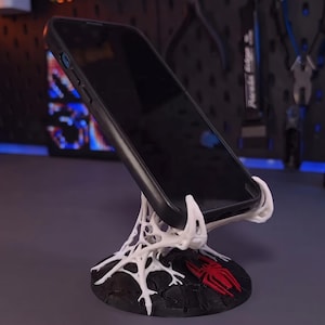 Peut inclure: Un smartphone noir posé sur un support imprimé en 3D blanc en forme de toile. Le support présente une base circulaire noire avec un emblème d'araignée rouge intégré à la surface. La structure blanche soutient le téléphone incliné.
