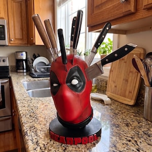 Può includere: Un ceppo portacoltelli a forma di testa del personaggio Deadpool, rosso e nero, con fori bianchi per gli occhi, poggiante su una base circolare nera con la parola DEADPOOL in lettere rosse in rilievo. Contiene sei coltelli da cucina.