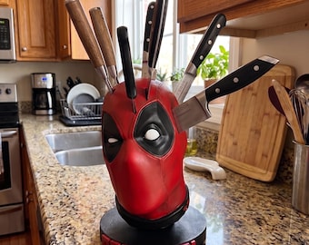 May include: A red and black Deadpool character head knife block featuring white eye cutouts, resting on a circular black base with the word DEADPOOL in raised red letters. The block holds six various kitchen knives with wooden and black handles.