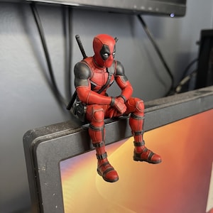 May include: A Deadpool action figure in a red and black suit with a mask, white eye patches, and black tactical gear. The figure is posed in a seated position with hands clasped, featuring a katana handle visible behind the shoulder.