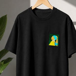 Op de afbeelding: Een zwart T-shirt met korte mouwen en een vierkante patch op de linkerborst. De patch toont een gestileerde afbeelding van een persoon in profiel die een geel kledingstuk, een blauwe hoofddoek en een witte oorbel draagt tegen een donkergroene achtergrond.