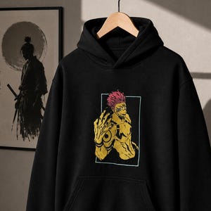 May include: A black hooded sweatshirt featuring a central graphic print of a yellow-skinned character with pink hair, dark markings, and a hand raised in a gesture. The hoodie includes a front kangaroo pocket, long sleeves, and ribbed cuffs.