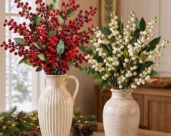 May include: Two ceramic vases holding artificial berry stems. The left vase is cream-colored with vertical ridges and a handle, filled with red berries and green leaves. The right vase is speckled beige, filled with white berries and green leaves.