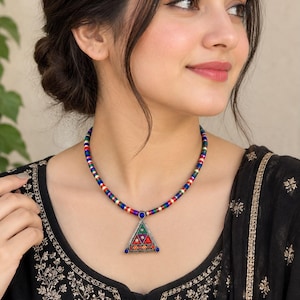 May include: A necklace featuring a multi-colored beaded cord in red, blue, green, and gold tones. The pendant is a silver-toned triangle shape with inlaid red, green, and blue stones, accented by small blue beads at each corner of the frame.