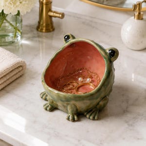 May include: A ceramic frog-shaped trinket dish with a glossy green exterior and a smooth pink interior. The bowl features two black eyes on the rim and four webbed feet at the base. Inside the dish are gold-toned necklaces and rings.