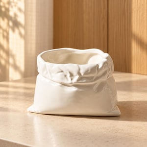 May include: A white ceramic vessel shaped like a crumpled paper bag. The container features a glossy finish with realistic folds and creases along the top rim and sides, mimicking the appearance of a folded paper sack in a solid, rigid material.