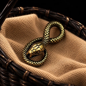 Puede incluir: Una figura de serpiente de metal en tono dorado enrollada en forma circular. La serpiente presenta escamas texturizadas detalladas en todo el cuerpo, una cabeza definida con ojos visibles y una boca ligeramente abierta, sobre una tela beige.
