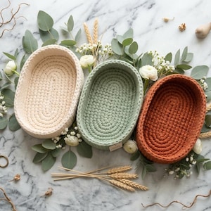 May include: Three oval-shaped baskets made of thick, crocheted cord in cream, sage green, and terracotta colours. Each basket features a consistent, textured stitch pattern and a deep, open interior, arranged together on a flat surface.