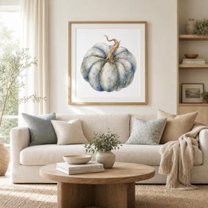 Op de afbeelding: Een vierkante ingelijste kunstdruk met een aquarelillustratie van een enkele blauwgrijze pompoen met een bruine steel, weergegeven in een lichthouten lijst gecentreerd aan een muur boven een beige bank met diverse decoratieve kussens.