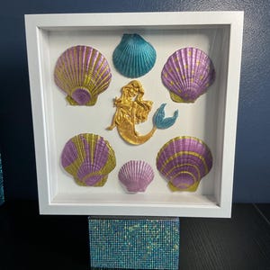 May include: A white shadow box frame containing a gold mermaid figure surrounded by five painted seashells. The shells feature metallic purple, gold, and teal colours. The frame rests on a rectangular base covered in small, iridescent blue rhinestones.