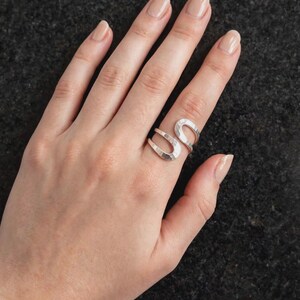 Può includere: Un anello aperto color argento con una finitura martellata e testurizzata. La fascia è formata da due distinte sezioni curve a forma di U che si incontrano al centro, creando un design a bypass che avvolge il dito con una lucentezza metallica.