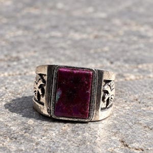 Puede incluir: Un anillo de metal plateado con una piedra rectangular de color púrpura intenso engastada en el centro. La banda tiene un acabado texturizado y desgastado, e incluye diseños de palmeras en relieve a ambos lados del engaste central.