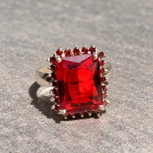 Pode incluir: Um anel de prata com uma grande pedra preciosa vermelha retangular e facetada como peça central. A pedra central é rodeada por um halo de pedras mais pequenas, redondas e de cor vermelho-escura, cravadas em prata, criando um aspeto texturado.