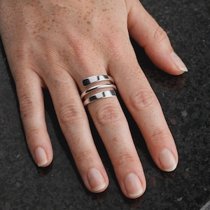 May include: A polished silver ring featuring a triple-band design. The ring consists of three smooth, interconnected metallic bands that wrap around the finger, creating a layered appearance with a reflective, high-shine finish throughout.