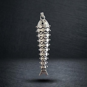 May include: A silver-toned metal pendant shaped like a fish, featuring a detailed scale pattern across its elongated body, a small circular eye, and a bail at the top for attachment. The surface has a polished finish with dark recessed lines.