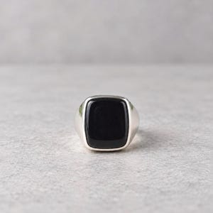May include: A silver signet ring featuring a smooth, polished, rectangular black onyx stone set into the center. The band is made of a solid, reflective silver-toned metal with a clean, rounded profile that tapers toward the back of the ring.