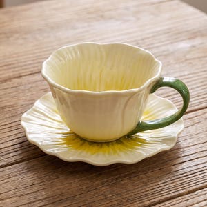 May include: A ceramic teacup and saucer set shaped like a flower. The cup has pale yellow, scalloped, petal-like walls and a smooth, curved green handle. The matching saucer features a scalloped edge with a yellow center and cream-colored petals.